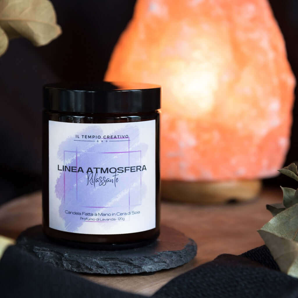 Candela profumata in cera di soia al profumo di lavanda per un'atmosfera rilassante, con una lampada di sale rosa dell'Himalaya sullo sfondo. Realizzata in vasetto contenitore di vetro ambrato.