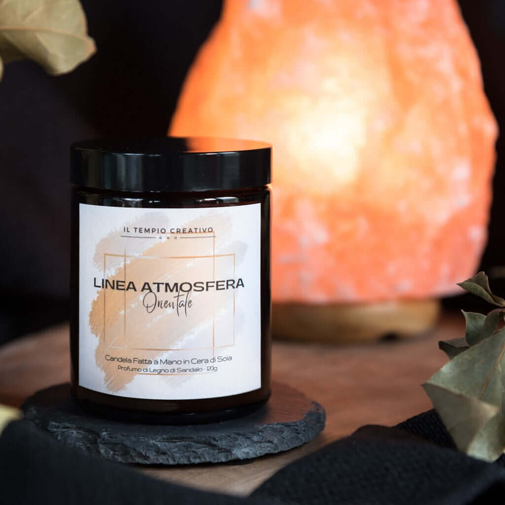 Candela profumata in cera di soia al profumo di legno di sandalo per un'atmosfera orientale, con una lampada di sale rosa dell'Himalaya sullo sfondo. Realizzata in vasetto contenitore di vetro ambrato.