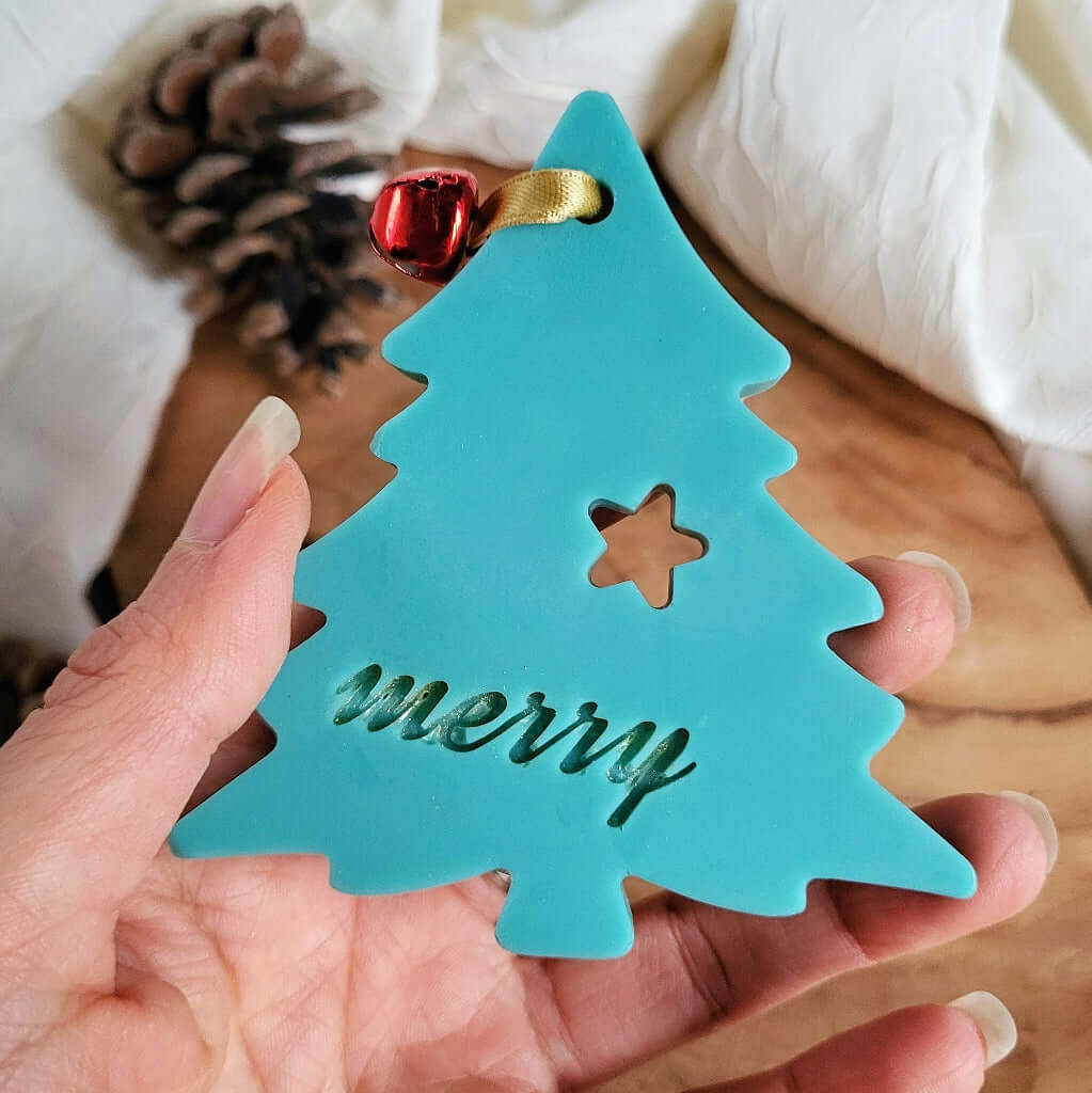 Tartina Profuma Armadi e Cassetti in cera di soia naturale al profumo di Pino. È a forma di Albero di Natale e Decorata con campanellino rosso. Scritta Merry di Merry Christmas.
