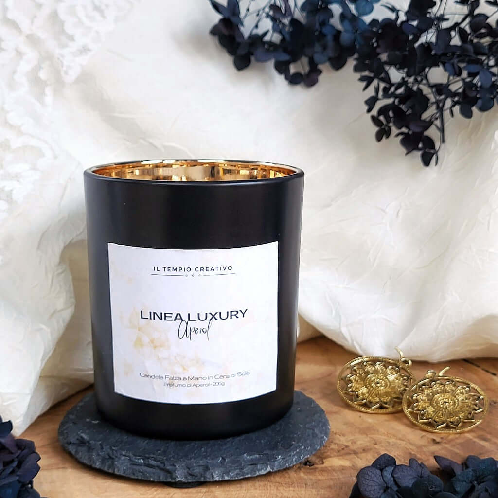 Candela profumata di lusso in cera di soia naturale. Realizzata in contenitore nero corvino e oro specchio con stoppino in legno. Design minimal e ricercato. Fragranza Aperol